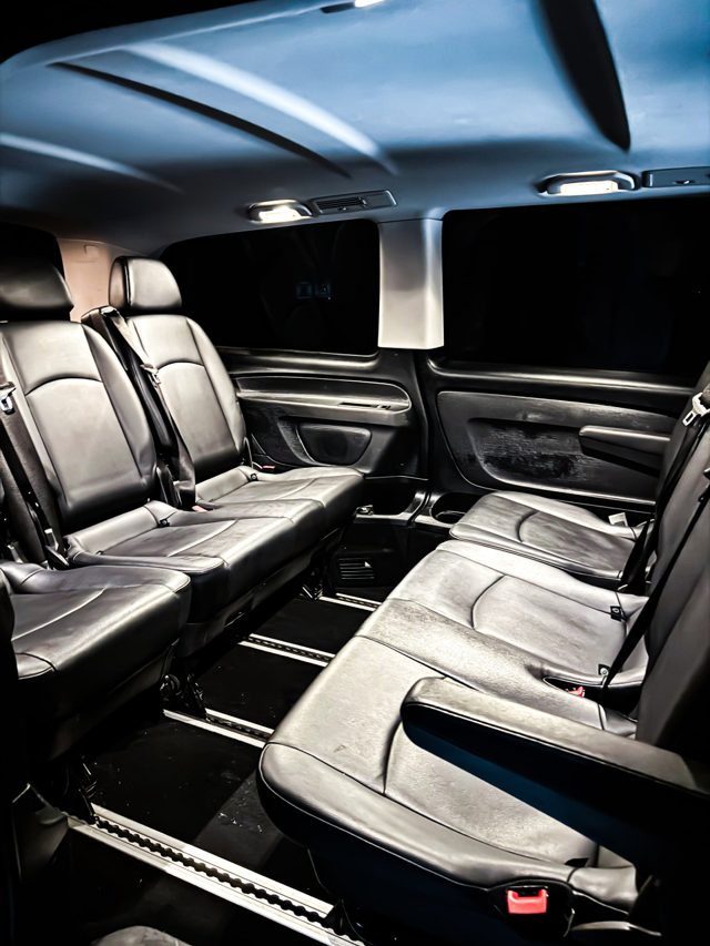 Rear passenger area of a van with black leather seats facing each other, lit by ceiling lights, with dark tinted windows