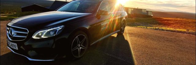Black Mercedes-Benz estate car parked on a tarmac road with low sun flaring over the roof, fell landscape and cabins behind