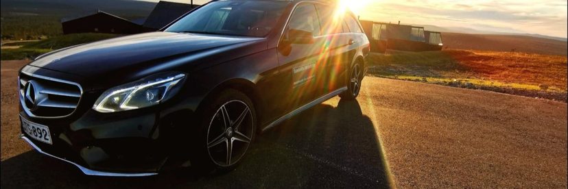 Black Mercedes-Benz estate car parked on a tarmac road with low sun flaring over the roof, fell landscape and cabins behind