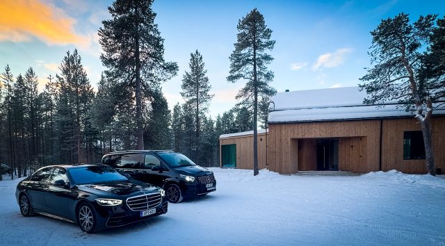 Black Mercedes-Benz sedan and van parked on snow beside a timber building among tall pine trees at dusk
