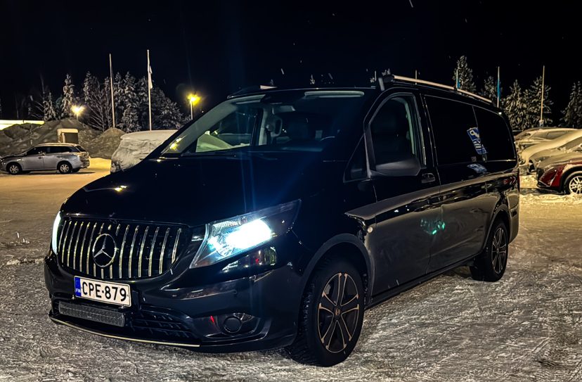 Black Mercedes-Benz van with headlights on, parked on packed snow at night with snowy trees and parked cars behind