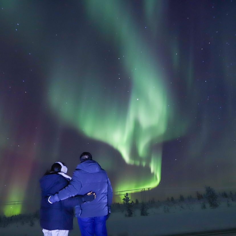 Two people in winter jackets stand arm in arm, watching bright green northern lights swirl above a snowy field at night
