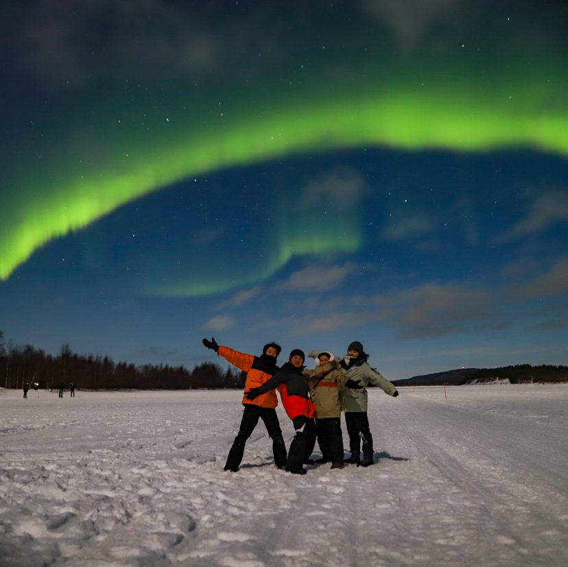 Four people in winter clothing pose with arms outstretched on a snowy open area under a bright green aurora arc
