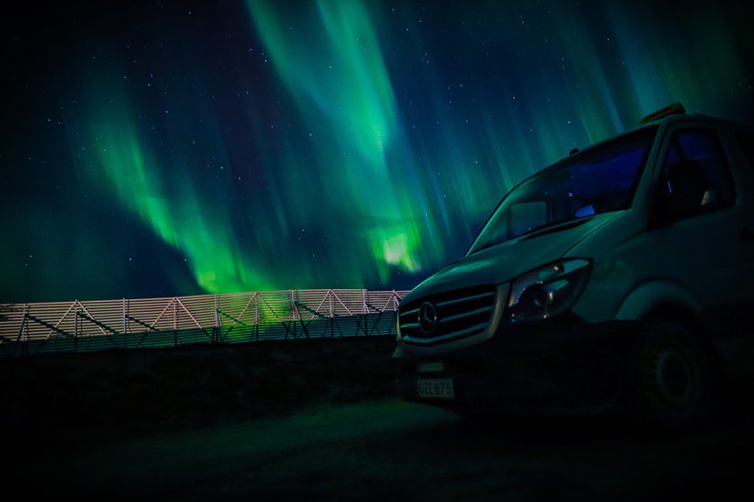 Mercedes-Benz van with a taxi roof sign parked at night beside a fence, with green northern lights across the starry sky