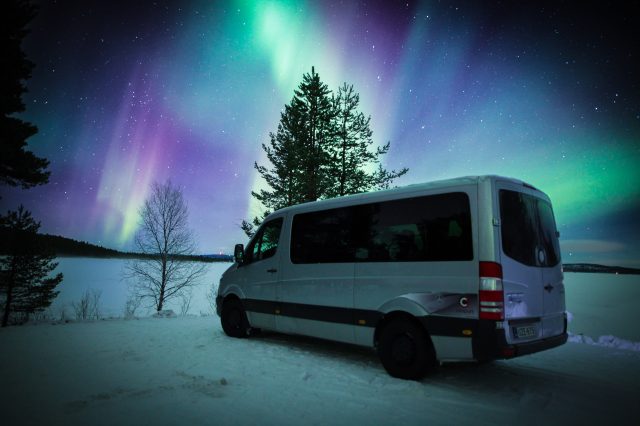 Silver Mercedes-Benz van parked on snow beside a frozen lake under green and purple northern lights and a starry sky
