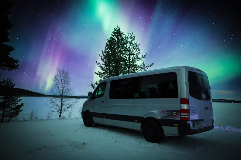 Silver Mercedes-Benz van parked on snow beside a frozen lake under green and purple northern lights and a starry sky