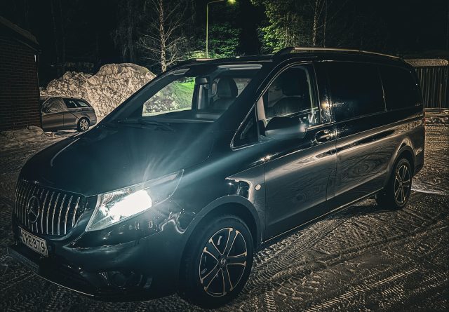 Dark Mercedes-Benz Vito van with headlights on, parked on a snowy surface at night beside snow piles and a parked car