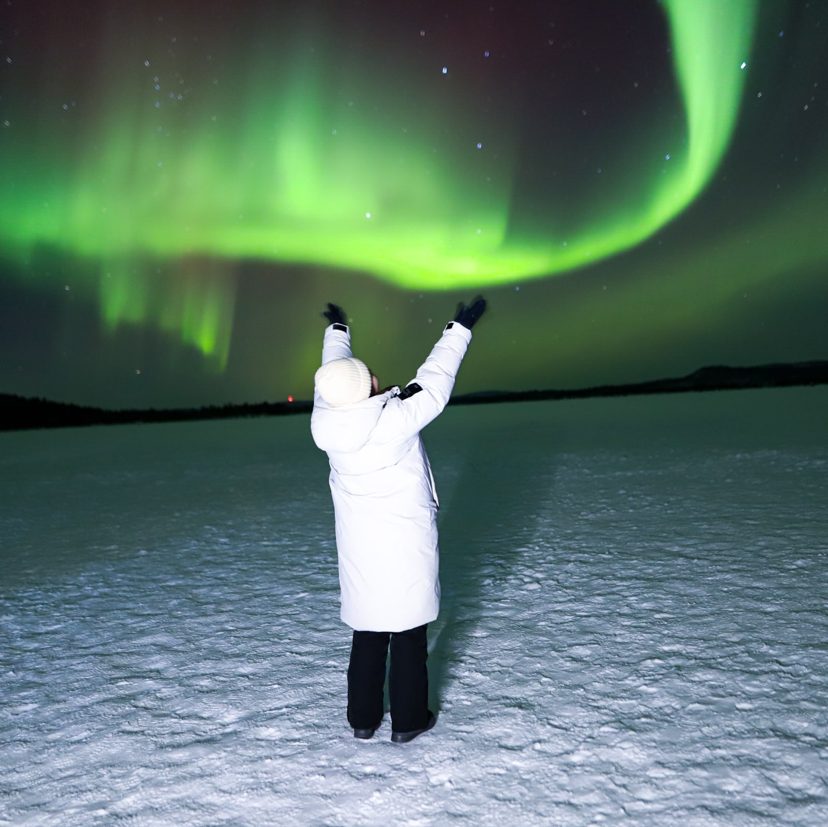 Person in a white winter coat and beanie raises both arms toward bright green northern lights over a snowy open expanse at night
