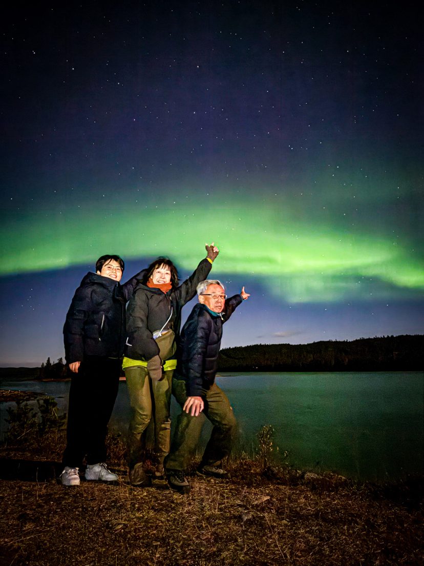 Three people in winter jackets pose and point upward beside a lake under a starry sky with green northern lights