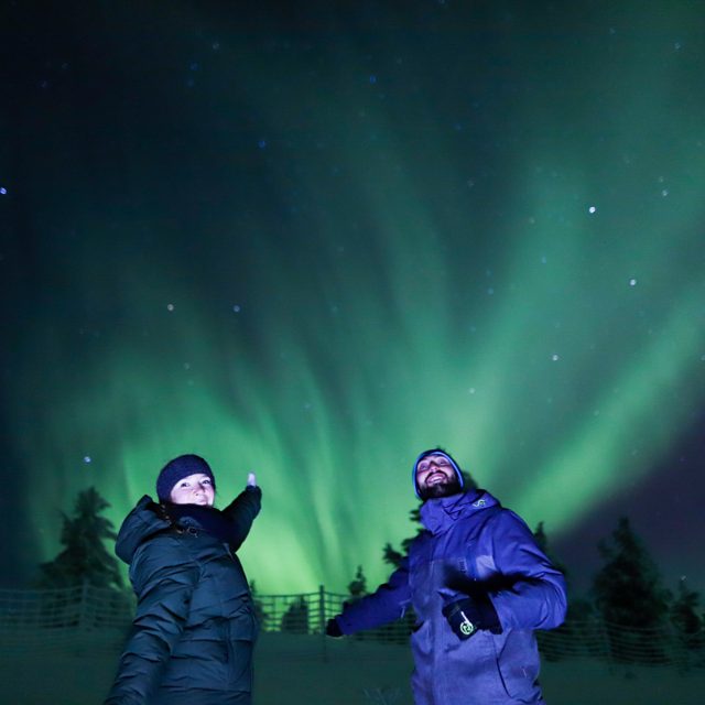 Two people in winter jackets and beanies pose under bright green northern lights and stars, with snow and fir trees behind