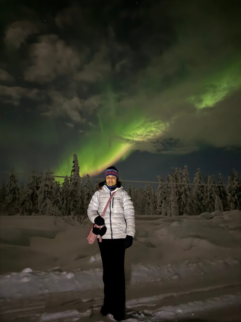 Woman in a white puffer jacket and beanie stands on snow at night in Tankavaara, with green northern lights over snowy spruce trees